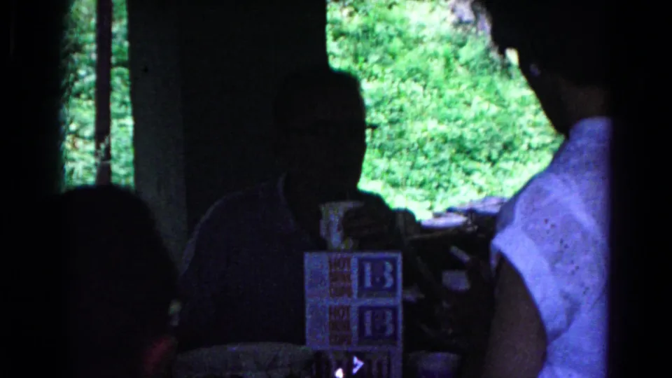 A dim 1963 home-movie frame shows adults in silhouette around a table, with a sleeveless white blouse, mugs, and boxed food visible.