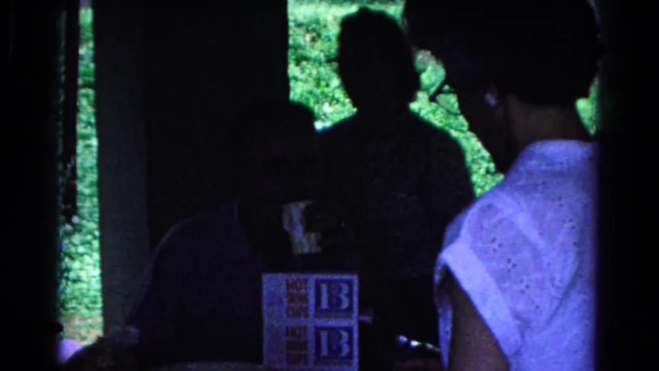 At the end of the clip, adults remain gathered in the shaded Cold Springs setting, with mugs, cigarettes, and HOT BUNS packaging on the table.