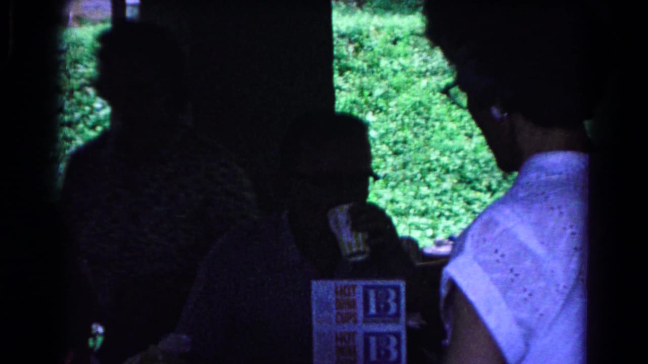 1963 Cold Springs home-movie frame showing silhouetted adults in a park shelter beside drink-cup cartons and sunlit trees.
