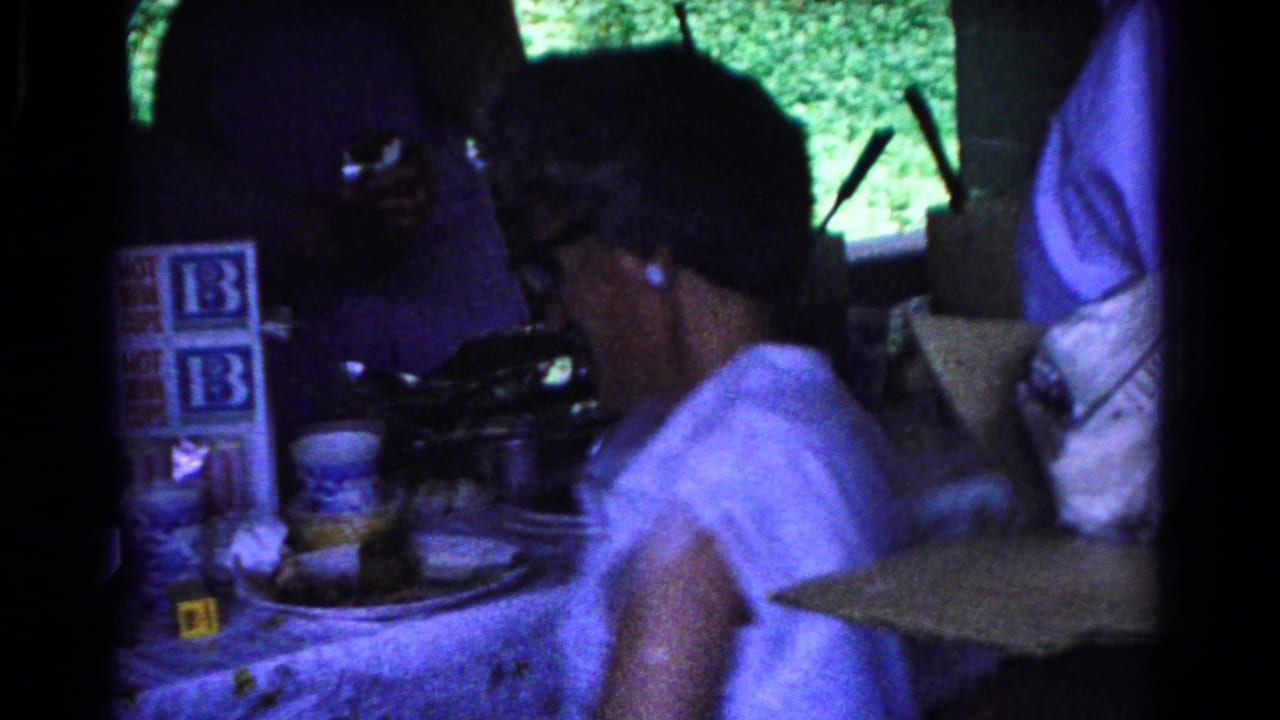 A person in a white top stands beside a table with cups and drink-cup cartons in a pavilion.