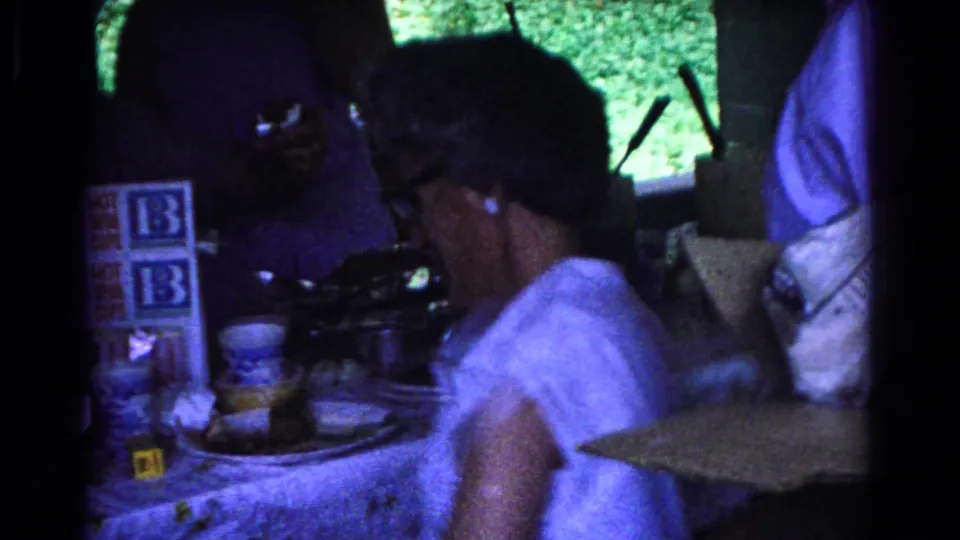 1963 Cold Springs park gathering frame with a person in a white patterned top near paper cups, cartons, and shaded pavilion posts.