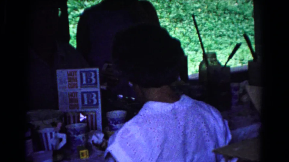 1963 Cold Springs home-movie view of a shaded pavilion table with drink-cup cartons, cups, and a person seen from behind.