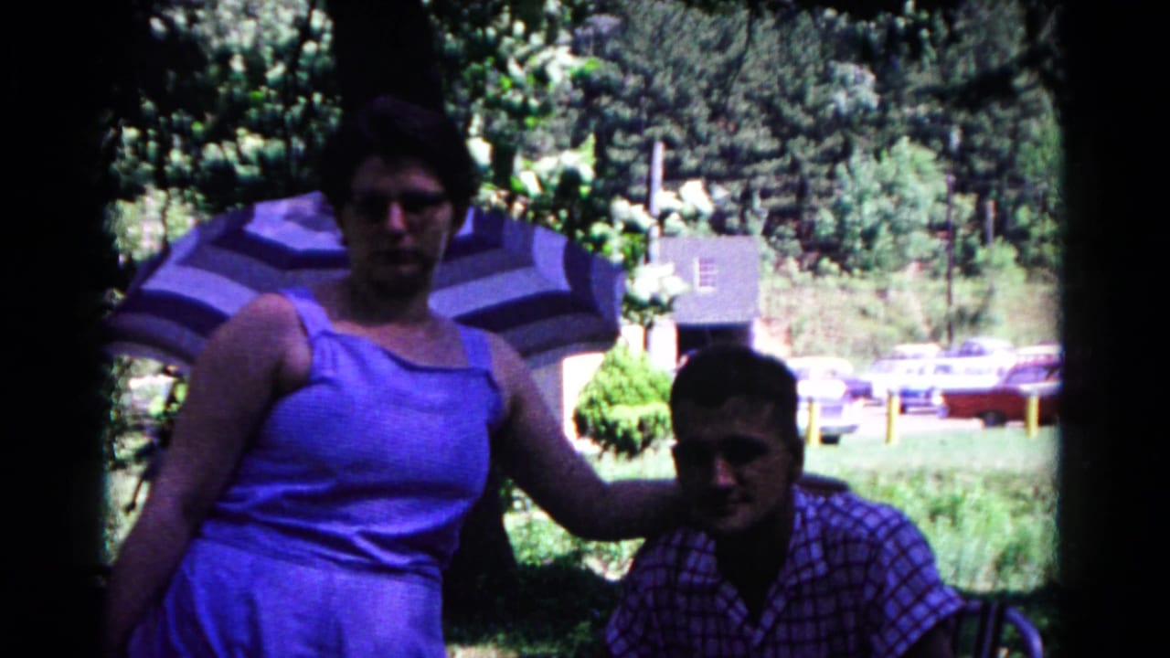 In supplied 1963 Cold Springs home-movie footage, a woman in a blue dress poses beside a seated man near trees, an umbrella, and parked cars.