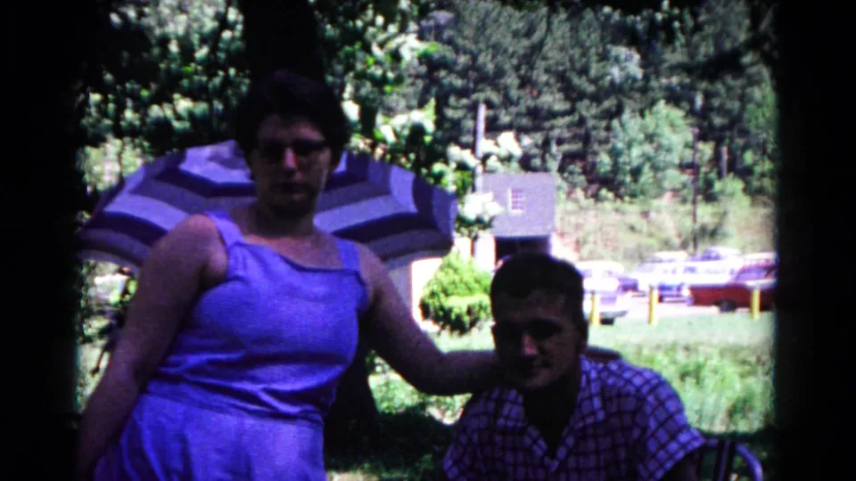 In supplied 1963 Cold Springs home-movie footage, a woman in a blue dress poses beside a seated man near trees, an umbrella, and parked cars.
