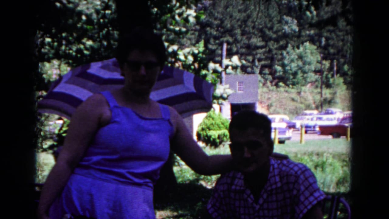 A woman in a blue dress stands beside a seated man in shaded park light, with a striped umbrella and cars visible in the background.