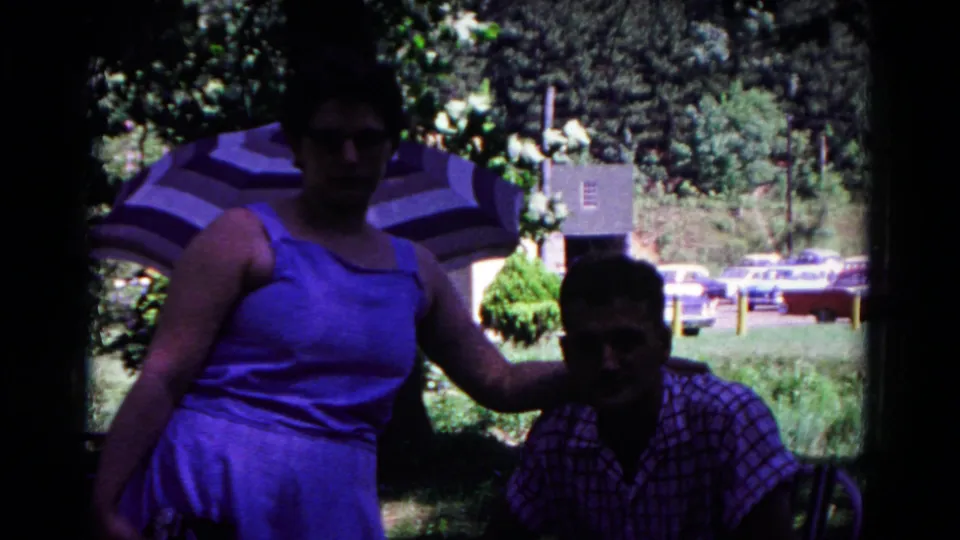 The home-movie frame holds on a woman beside a seated man at a park, backed by a sun umbrella, trees, grass, and mid-century cars.