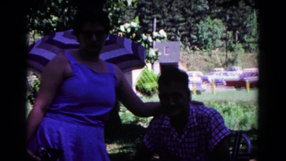 At the end of the short 8mm clip, the woman and seated man remain posed in a park setting with umbrella shade and parked cars behind them.