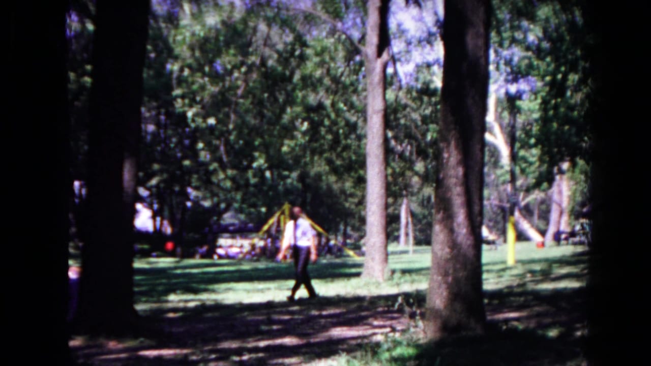 Man in a light shirt walks through a shaded park with trees and a playground behind him.