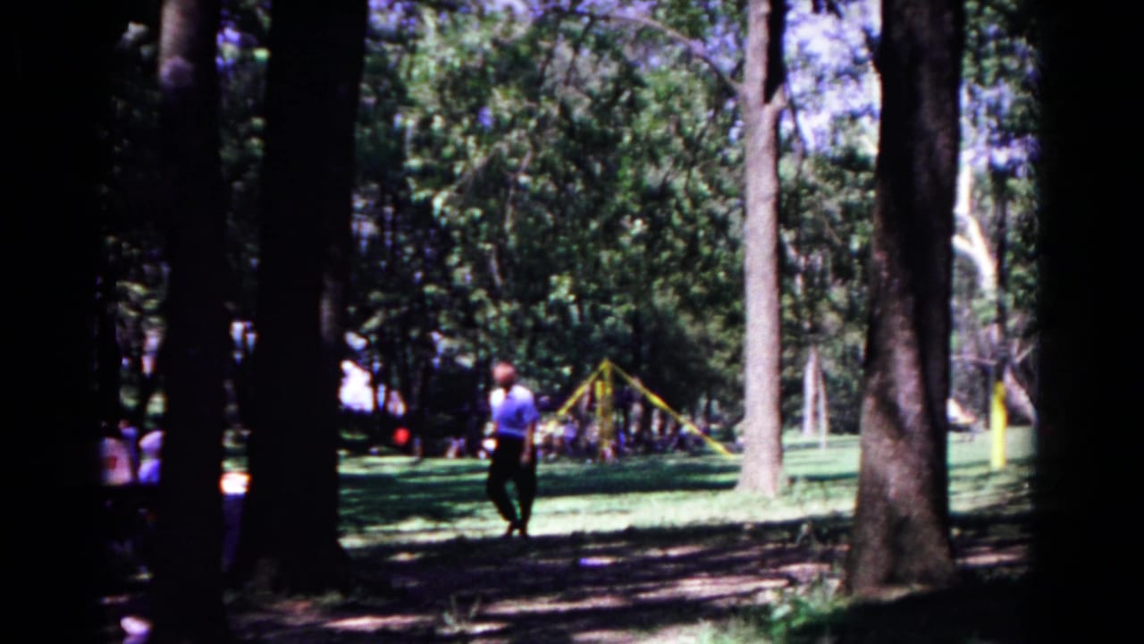 The man continues through the shaded Cold Springs park, framed by tall tree trunks and a sunlit playground area in the distance.