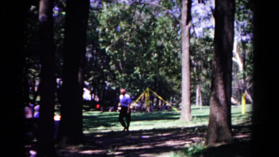 The man continues through the shaded Cold Springs park, framed by tall tree trunks and a sunlit playground area in the distance.