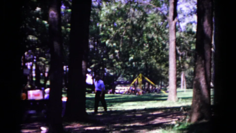 A 1963 home-movie view shows the man near the left side of the wooded park, with playground equipment and sunlit grass in the background.