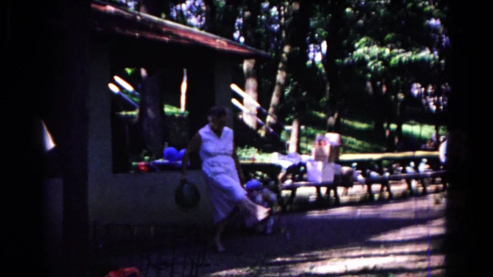 In Cold Springs, New York, 1963 home-movie footage, a woman in a white dress walks near a picnic shelter with balloons and benches nearby.