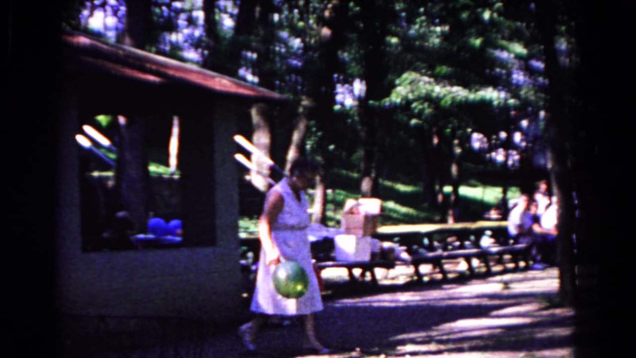 Woman carries a green balloon while walking past picnic tables in a shaded park.