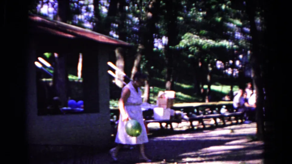 The woman walks through the Cold Springs park scene carrying a green balloon, with picnic benches and seated people behind her.
