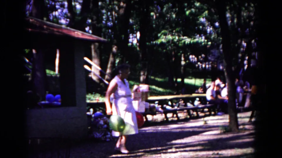 In the 1963 Cold Springs park footage, the woman pauses near the shelter with a green balloon as benches and trees fill the background.