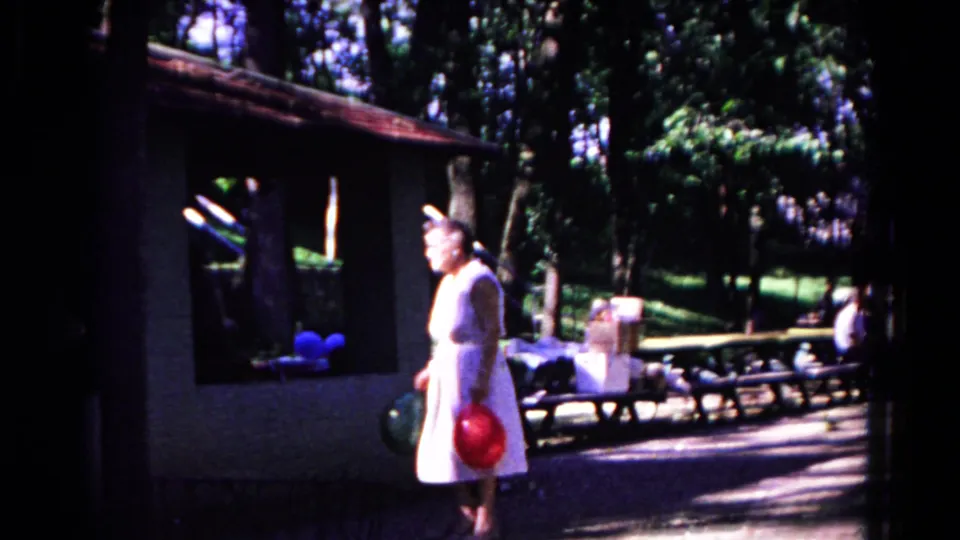 The woman stands in the Cold Springs park with red and green balloons, framed by a shelter, benches, trees, and seated visitors.