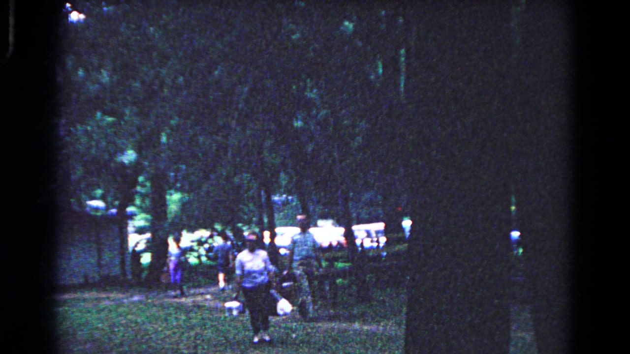 1963 home-movie frame in Cold Springs, New York, showing adults walking through trees toward a picnic area with baskets.