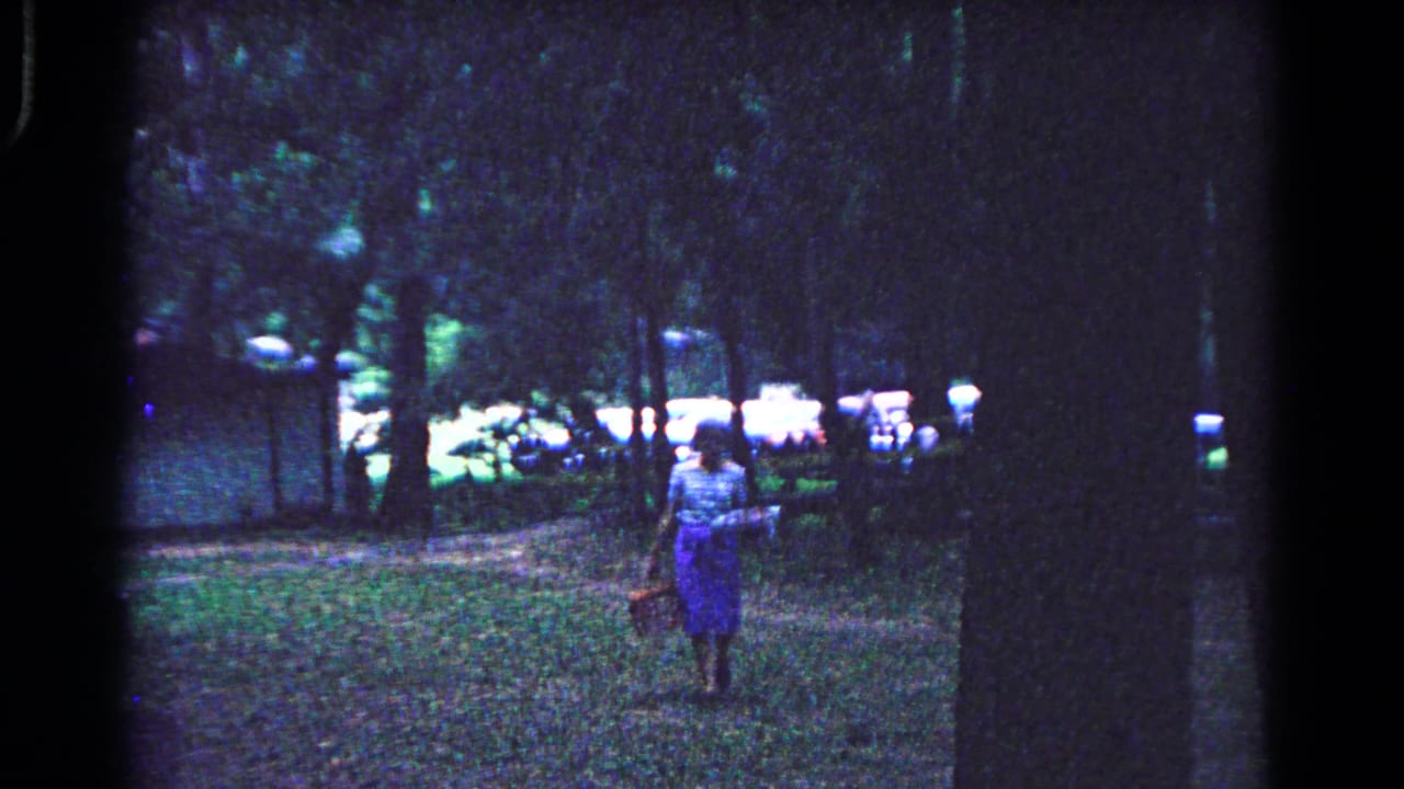Woman carrying a basket across a wooded campground clearing.