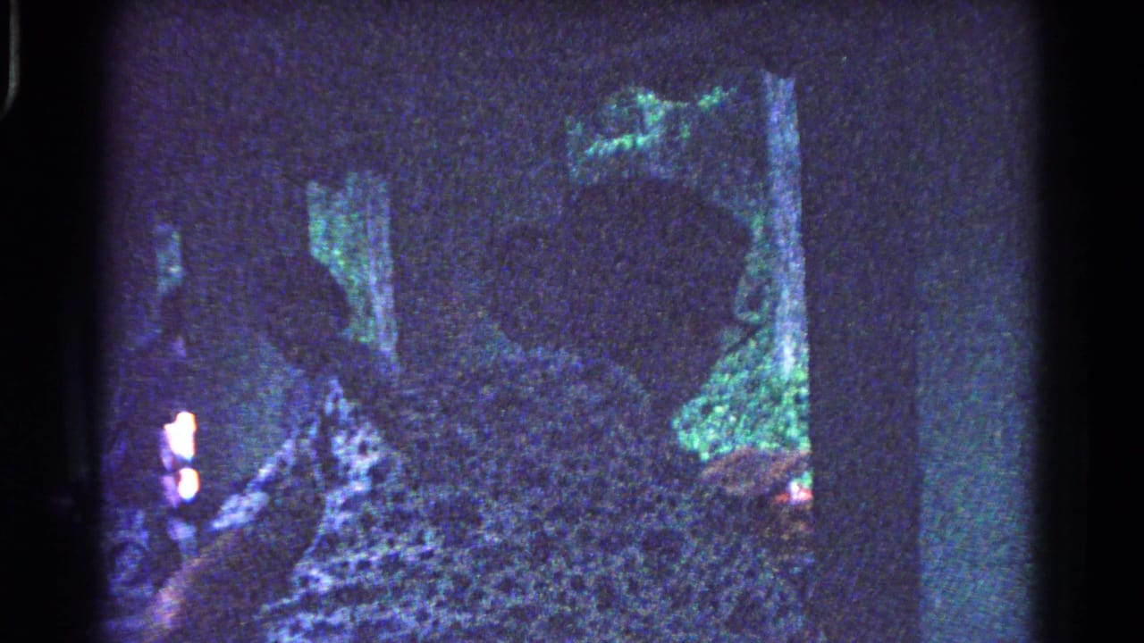 Silhouetted women arrange picnic items beneath a park shelter, seen through grainy blue-tinted 8mm home-movie footage.