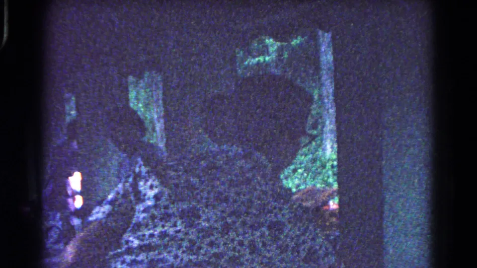 Silhouetted women arrange picnic items beneath a park shelter, seen through grainy blue-tinted 8mm home-movie footage.