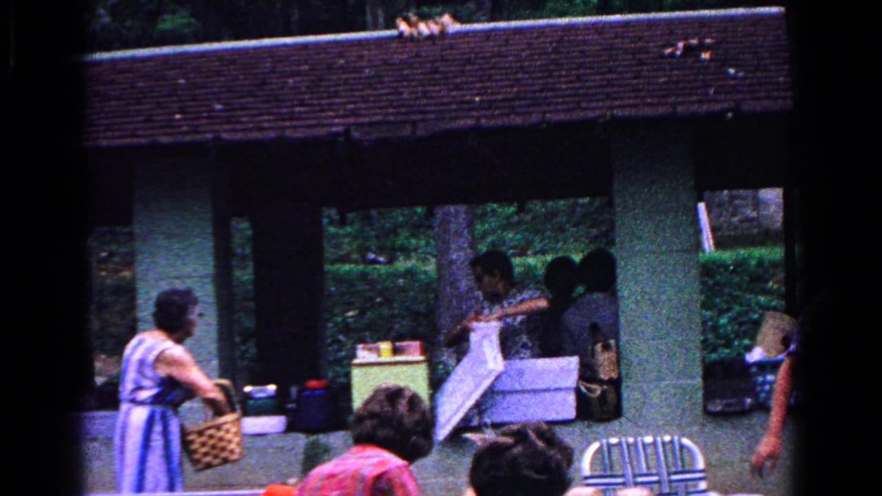 1963 Cold Springs home-movie frame of adults arranging picnic items under a wooden pavilion, with a striped dress, woven basket, coolers, and trees visible.