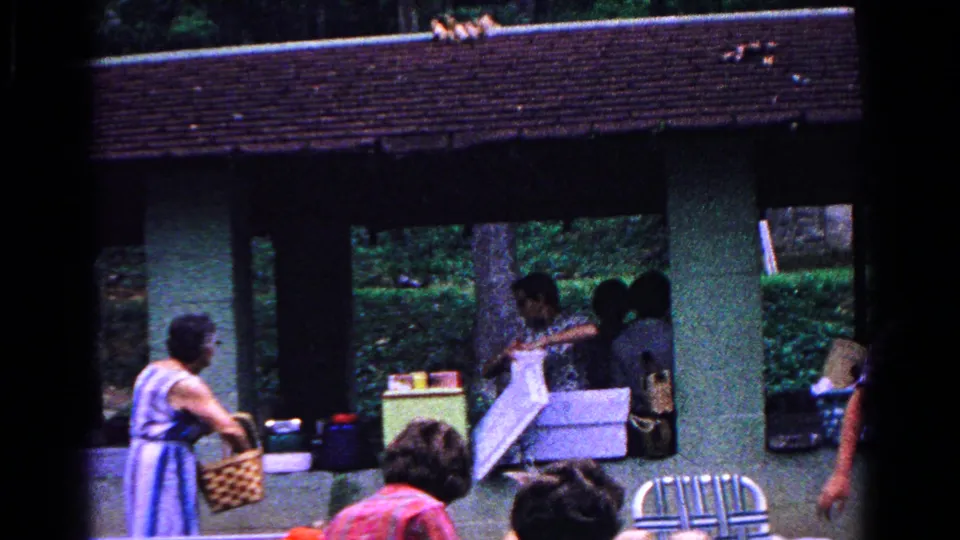 1963 Cold Springs home-movie frame of adults arranging picnic items under a wooden pavilion, with a striped dress, woven basket, coolers, and trees visible.