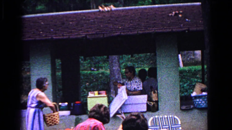 Cold Springs, New York, 1963 home-movie frame showing adults setting up a picnic with coolers, a basket, chairs, and wooded park background.