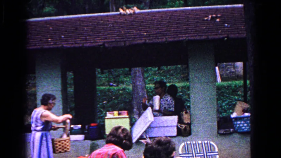 1963 Cold Springs family picnic frame with a woman leaning over a woven basket while others handle supplies under the shaded park pavilion.