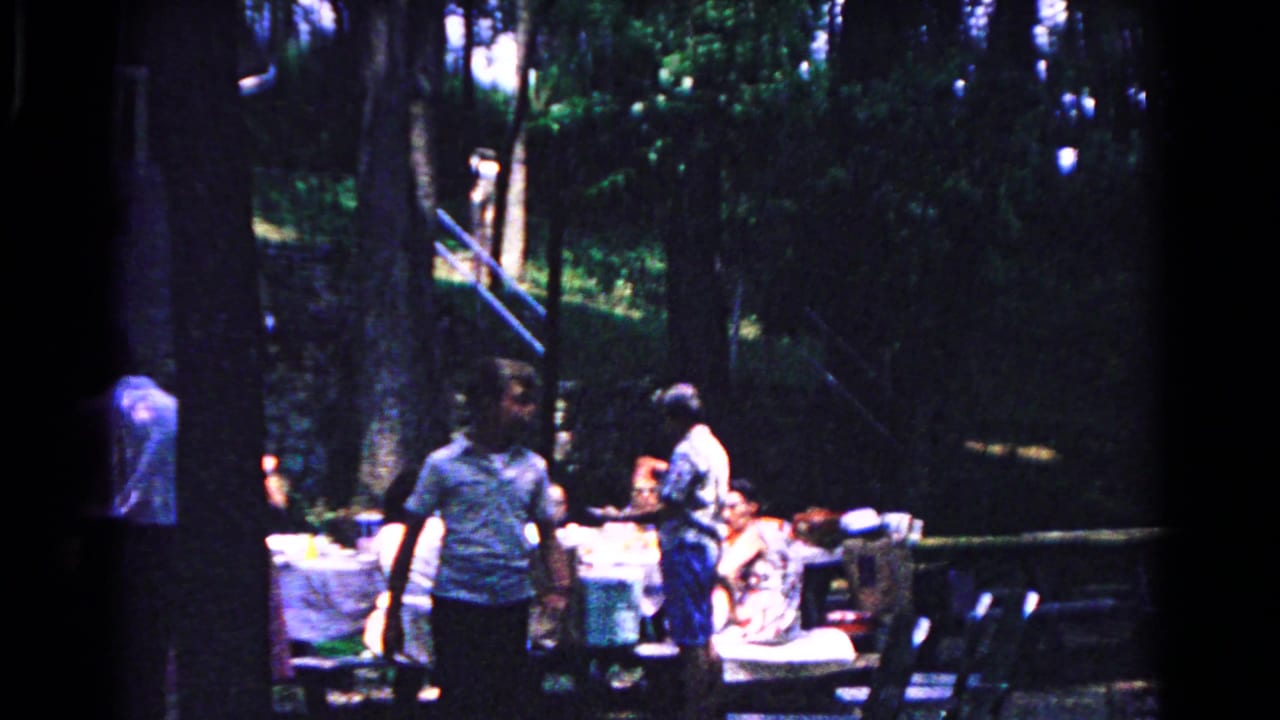 Adults gather around white-covered picnic tables in a wooded outdoor setting.