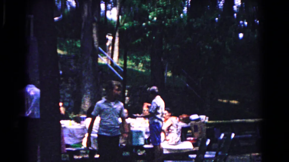 1963 home-movie frame from Cold Springs, New York, showing adults around picnic tables beneath tall trees.