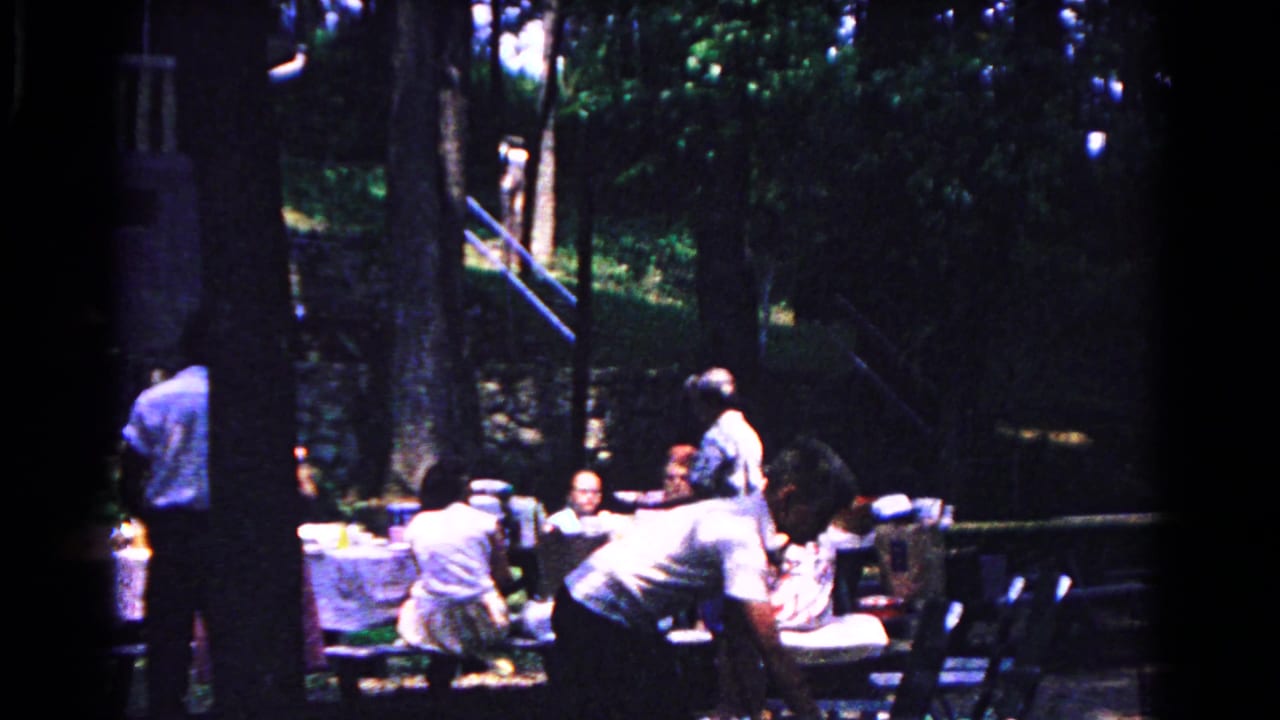 A wooded 1963 picnic scene in Cold Springs, New York, with adults seated and moving around tables.