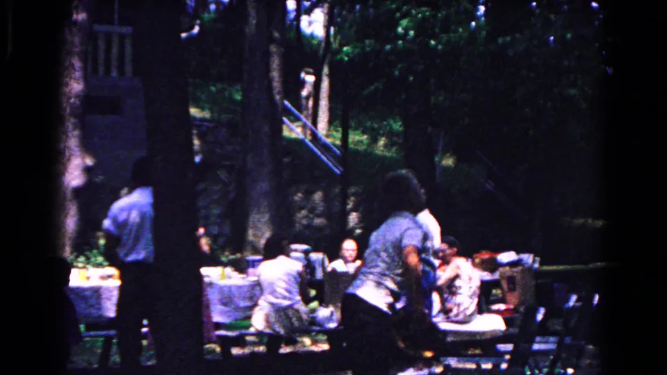 Cold Springs 1963 home-movie frame of adults gathered at a park picnic, with tables and trees visible.