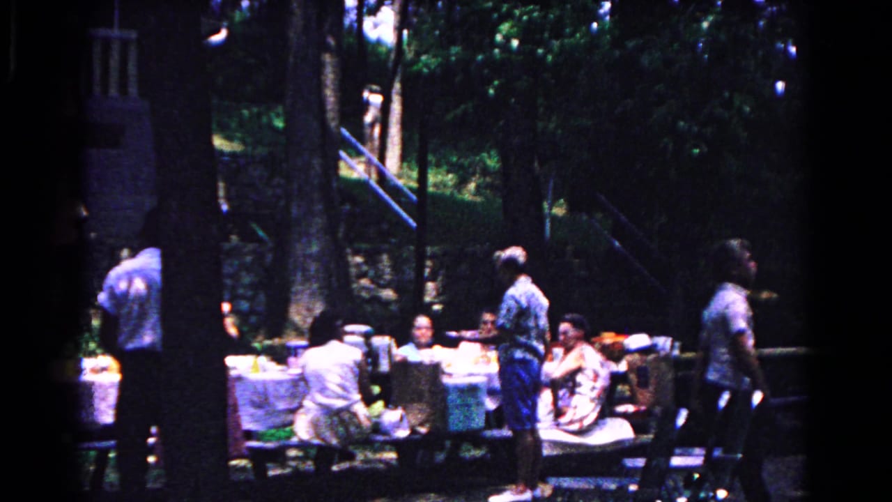 Adults gathered at picnic tables under tall trees in a wooded area.
