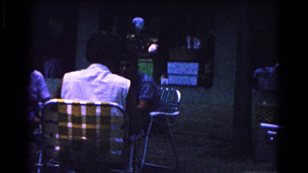 Dim 1963 Cold Springs home-movie view of adults seated near folding metal chairs beside a building or sheltered area.