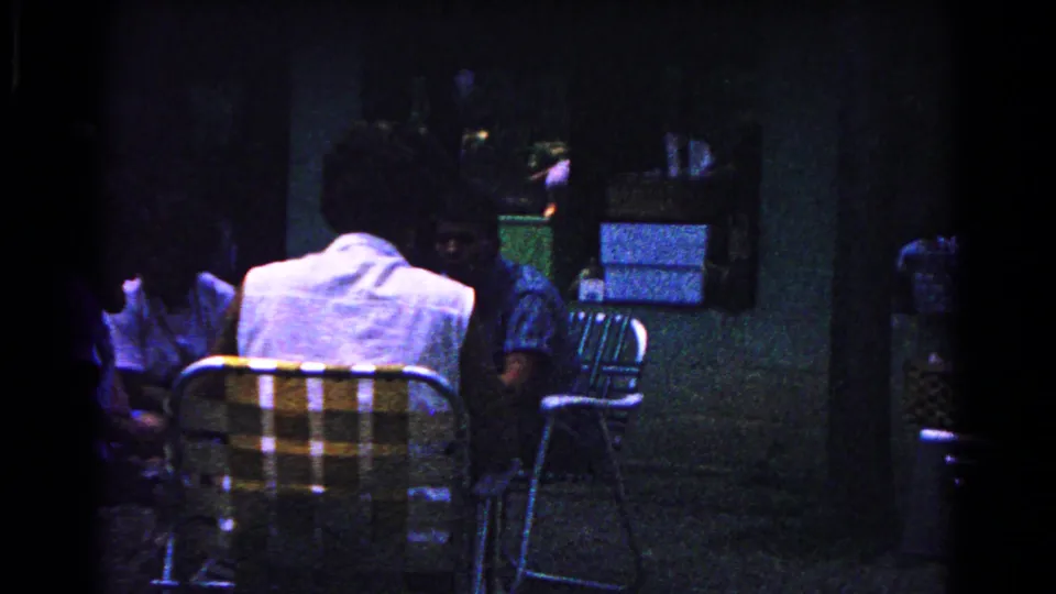 Low-light home-movie frame from the 1963 Cold Springs gathering, with adults seated behind a plaid folding chair.