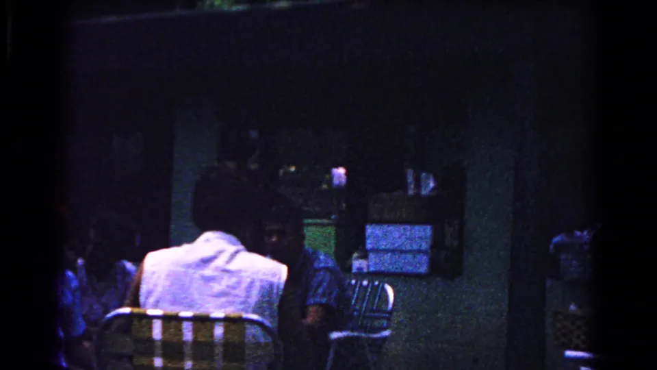 Dark 8mm frame from the Cold Springs family gathering, with seated adults, folding chairs, and a window visible in low light.