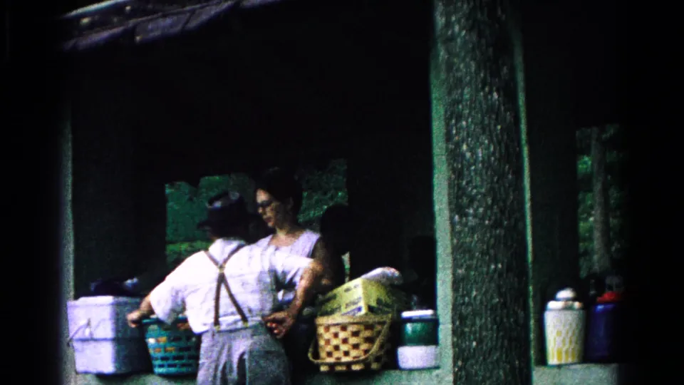 In Cold Springs, New York, 1963, two adults prepare picnic items under a shaded shelter with baskets, containers, and trees behind them.