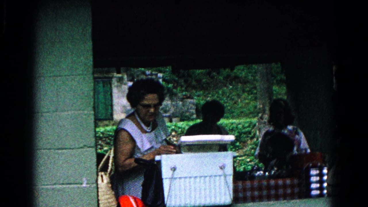 Woman looks down at a white cooler while other people sit near a picnic table.