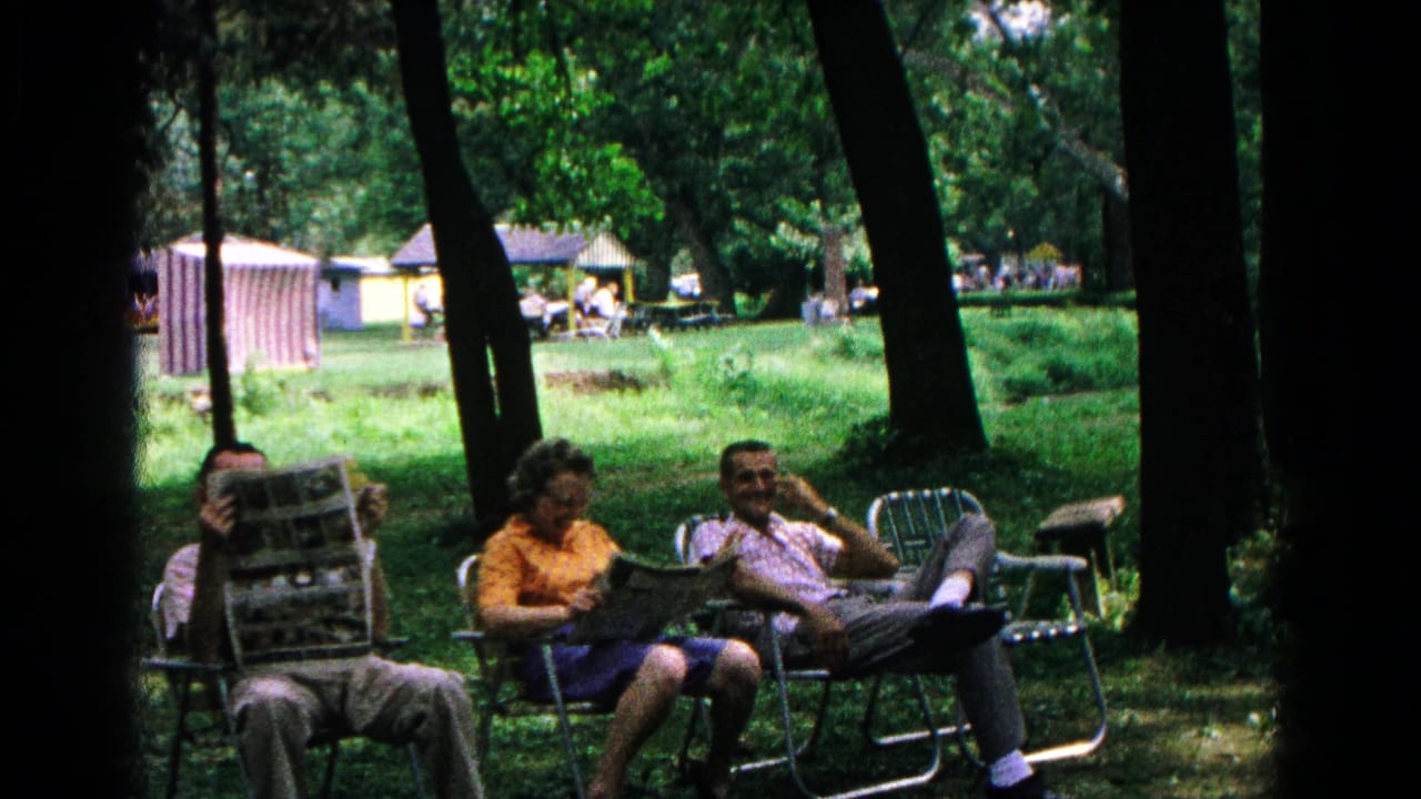 Adults sit in folding lounge chairs under trees, with one person holding a newspaper.
