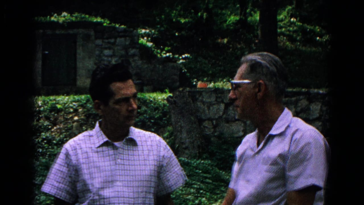 Two men appear to talk outdoors in Cold Springs, New York, with ivy, trees, and a low stone wall behind them.