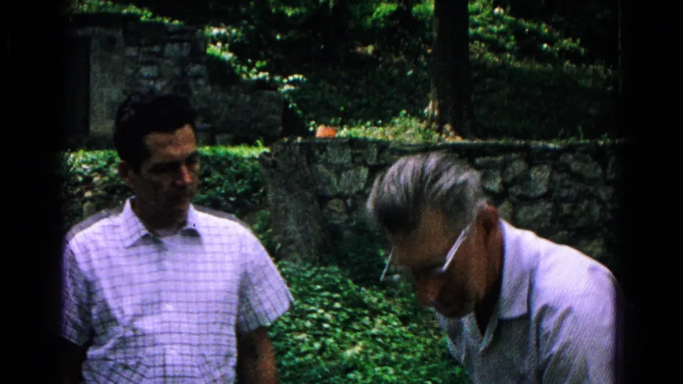 A younger man in a checked shirt stands as an older man with glasses bends forward near a stone wall in the outdoor setting.