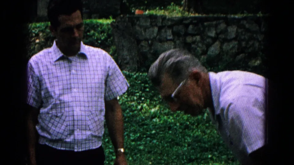 The outdoor home-movie frame shows one man standing and another bending forward against a backdrop of stonework and dense greenery.