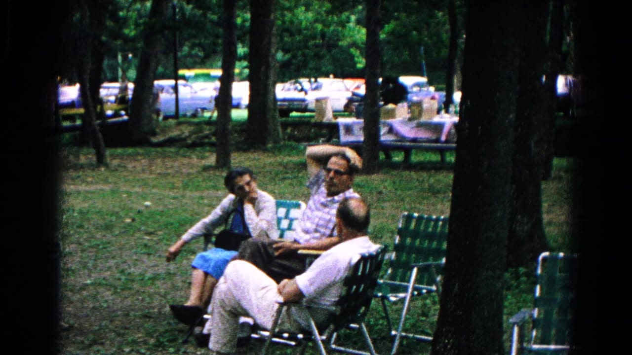 Three adults sitting in green lawn chairs under trees in a park.