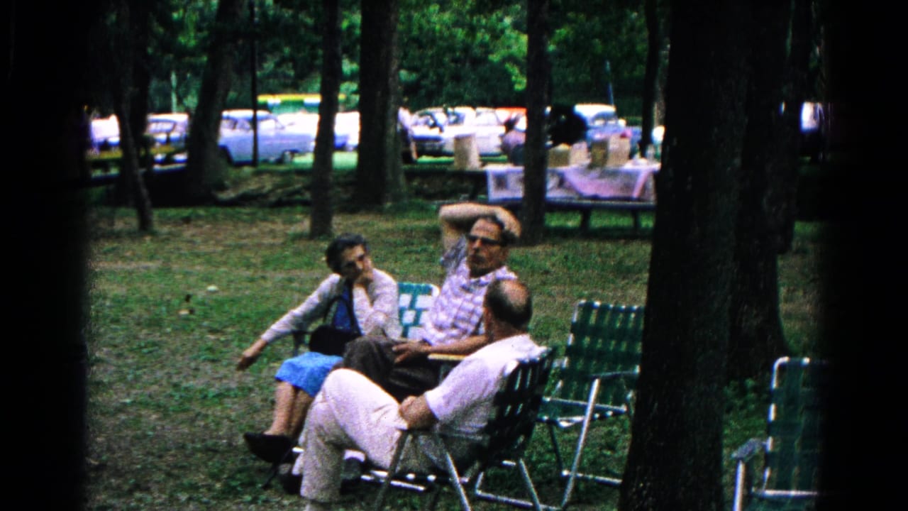 Adults seated in lawn chairs in a wooded park with cars behind them.