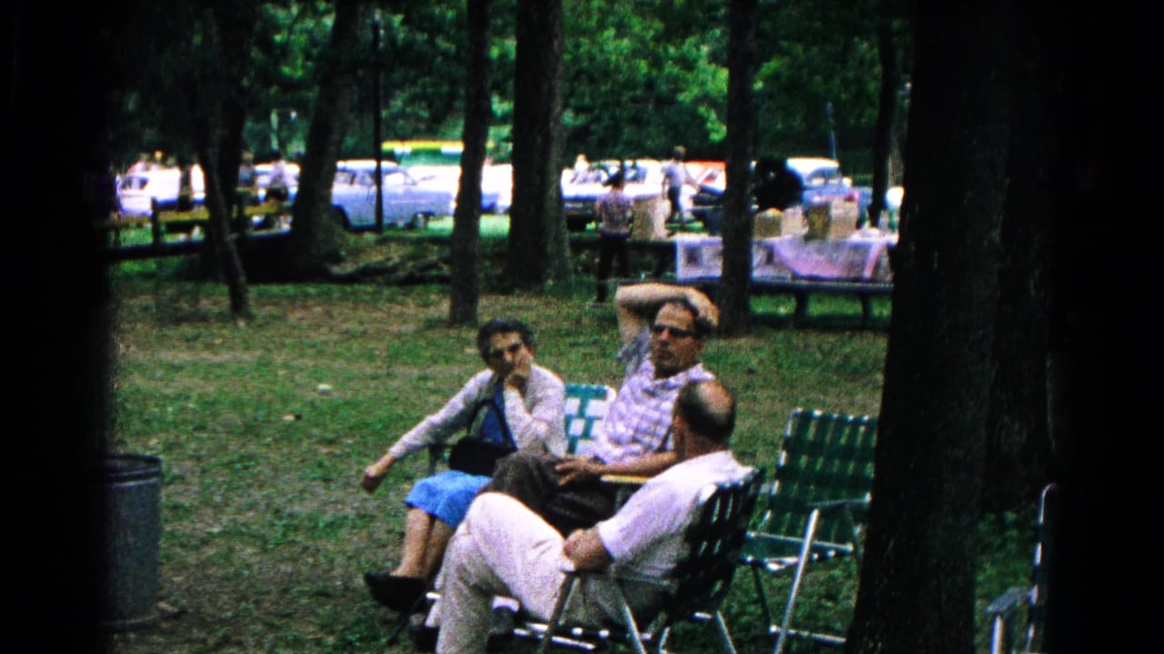 A home-movie frame shows three adults seated together in green webbed chairs in a wooded Cold Springs park setting.