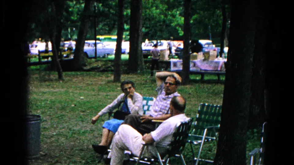 A home-movie frame shows three adults seated together in green webbed chairs in a wooded Cold Springs park setting.