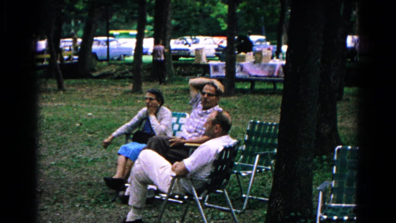 Three adults sit in green lawn chairs in a shaded park, with one man in the foreground and parked cars behind the tree line.