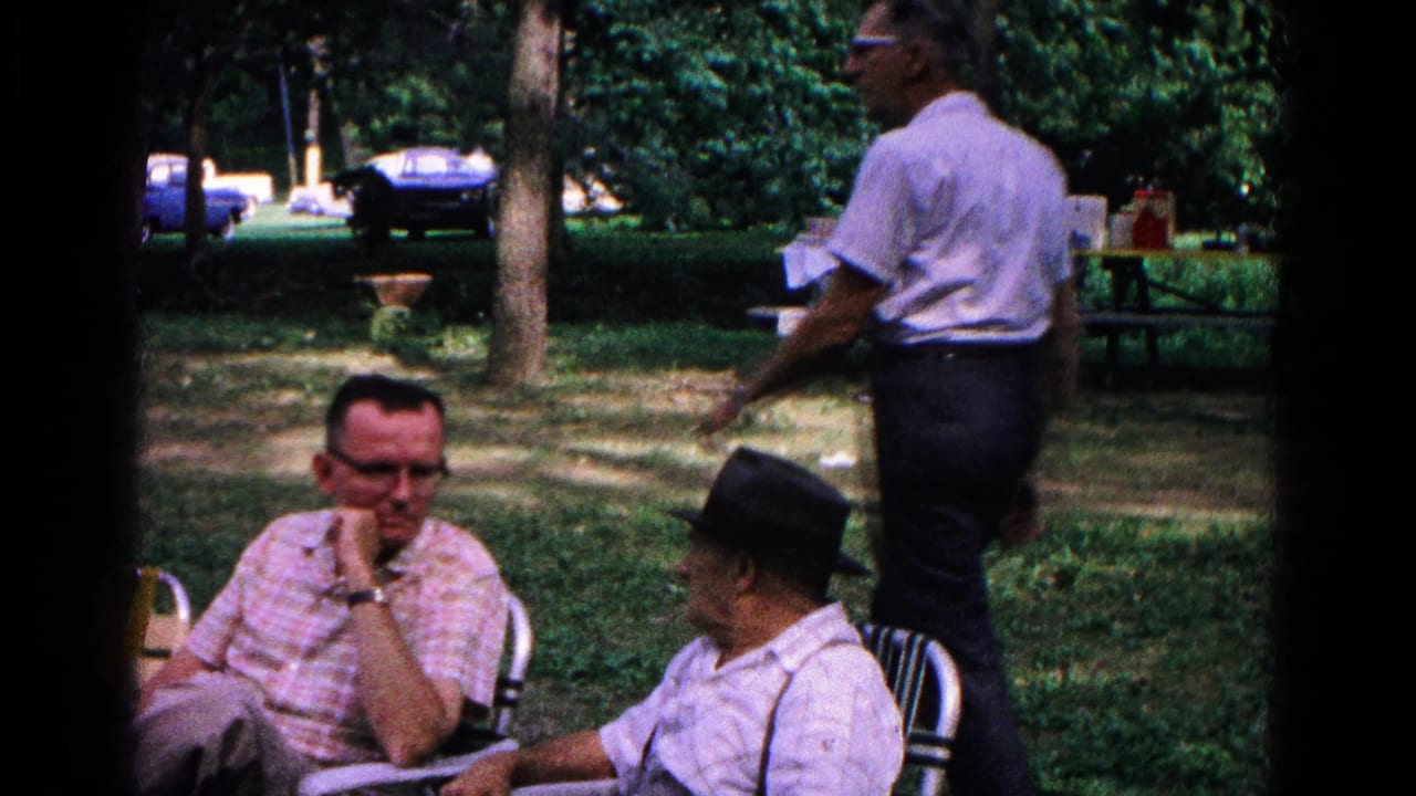 Adults seated in folding chairs as a man walks past in a wooded park.