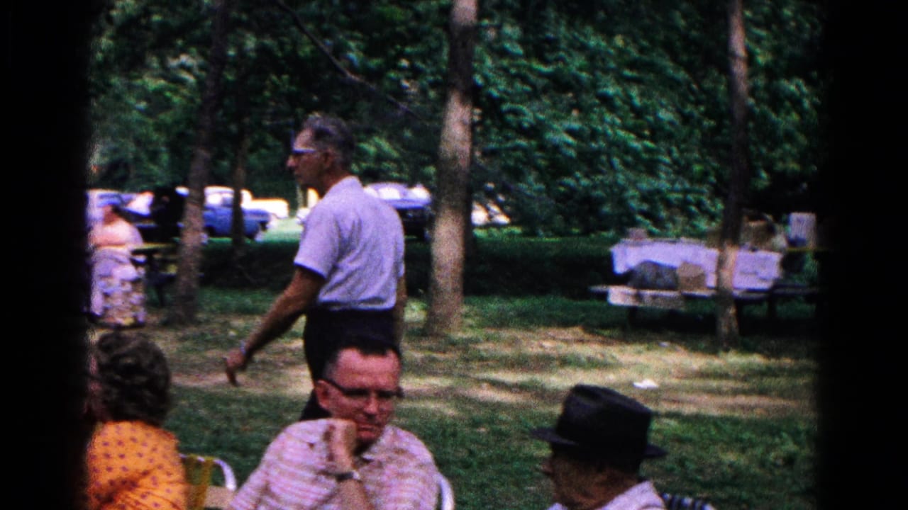A man walks behind seated adults in a wooded area with picnic tables and cars.