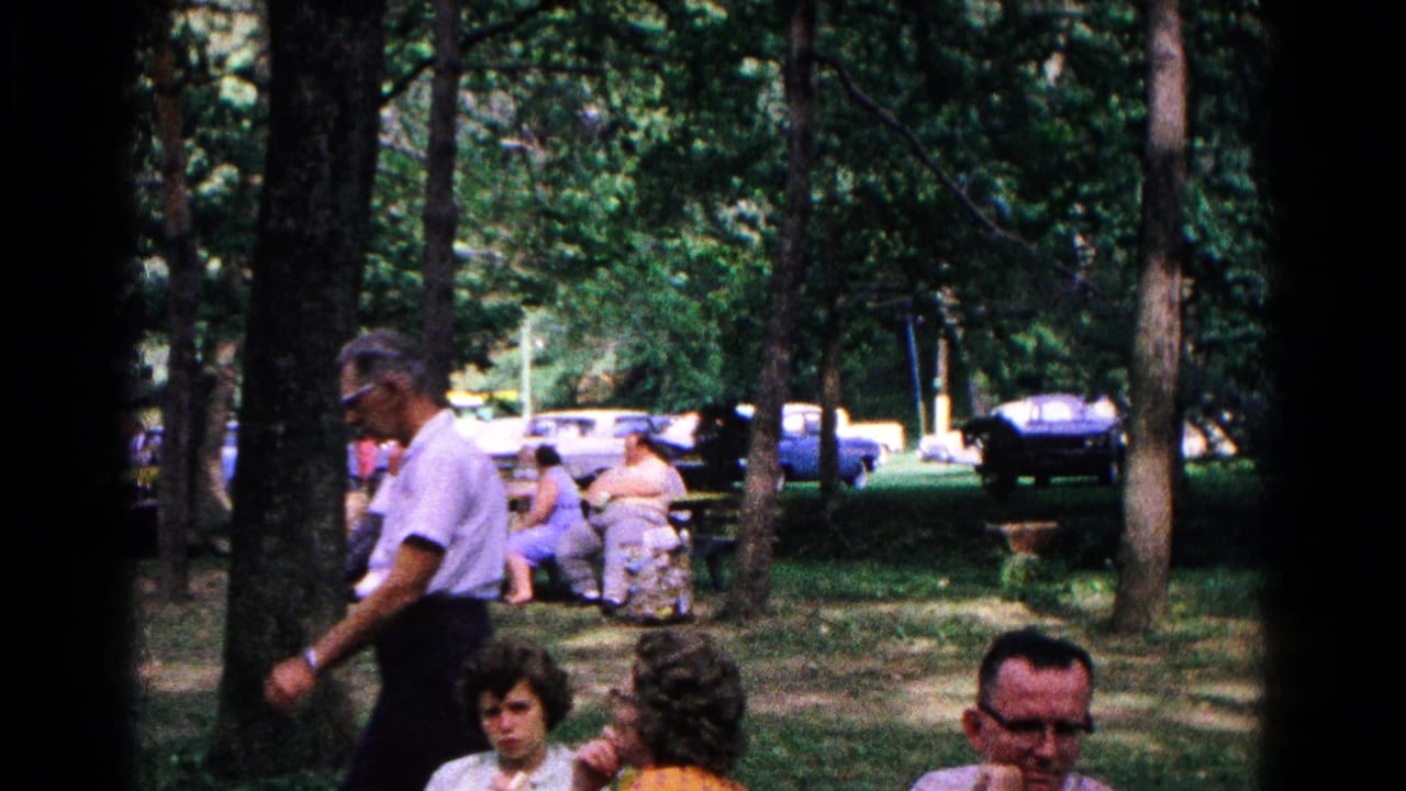 1963 Cold Springs home-movie frame showing adults seated in a shaded park as a man walks past trees with cars beyond.